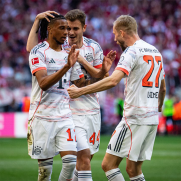 Trei jucători de fotbal în echipament alb și roșu sărbătoresc un gol. Unul dintre ei, cu părul lung, este în centrul atenției, zâmbind. Ceilalți doi îl îmbrățișează și îl felicită, creând o atmosferă de bucurie pe teren.