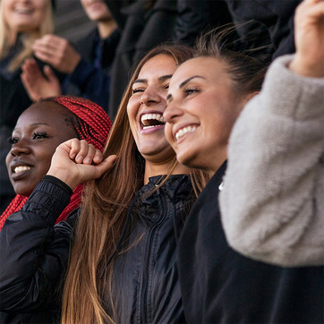 Trei femei zâmbitoare se bucură și strigă de bucurie, purtând haine de stadion. Femeia din mijloc are părul lung, iar celelalte au stiluri diferite de coafuri. Fundalul sugerează o atmosferă de entuziasm și celebrare.