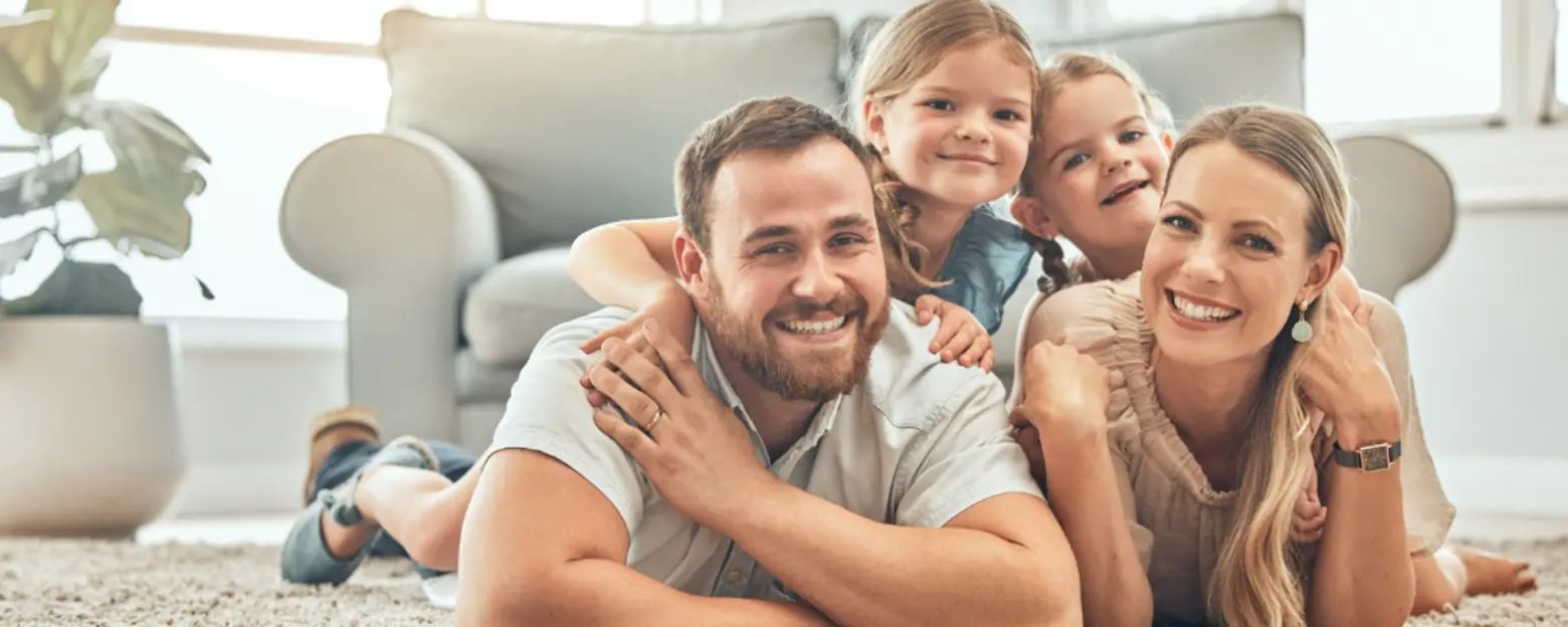 O familie fericită stă întinsă pe covor, zâmbind. Tatăl îmbrățișează copiii, iar mama se sprijină pe el, toți fiind relaxați și jucăuși. Fundalul sugerează un spațiu luminos și primitor, cu plante decorative și mobilă confortabilă.
