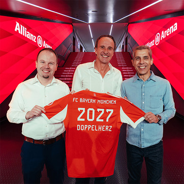 Trei bărbați stau în fața unei pereți roșii cu logo-ul Allianz Arena, ținând o tricou FC Bayern München. Tricoul are inscripționată cifra „2027” și cuvântul „DOPPELHERZ”. Aceștia zâmbesc, celebrând un moment important legat de echipă.