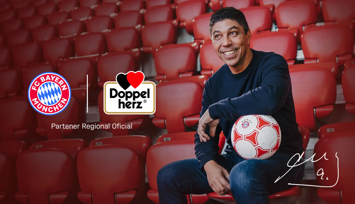 Un bărbat zâmbitor stă pe un scaun dintr-un stadion de fotbal, ținând în brațe o minge de fotbal. Între el și un logo aparținând echipei FC Bayern München și unul pentru Doppel herz, care menționează parteneriatul regional oficial.