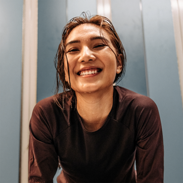 O tânără zâmbește cu bucurie, având părul ud, îmbrăcată într-o bluză de sport de culoare închisă, într-un mediu interior cu pereți albaștri. Expresia ei radiantă sugerează entuziasm și satisfacție.